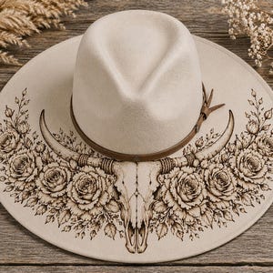 May include: A cream-colored wide-brimmed fedora hat featuring a brown leather band and a detailed dark brown illustration on the brim. The artwork depicts a central steer skull flanked by symmetrical arrangements of blooming roses and leaves.