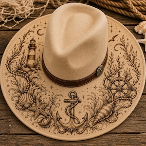 May include: A beige fedora hat with a brown leather band and a decorative button. The hat's brim features a nautical design with an anchor, seashells, a lighthouse, and a ship's wheel. The hat is set on a wooden surface.