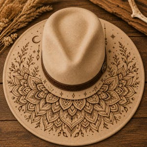 Puede incluir: Sombrero fedora beige con una banda de cuero marrón y un ala decorativa. El ala presenta un diseño de mandala detallado con acentos celestiales. Un accesorio elegante para diversas ocasiones.