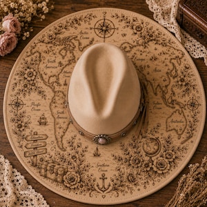 Puede incluir: Un sombrero fedora beige con una banda decorativa y un diseño de mapa del mundo en el ala. El ala presenta una rosa de los vientos, adornos florales y las palabras "Adventure, Freedom, Wild Heart."