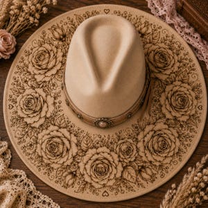 May include: A beige fedora-style hat with a wide brim, adorned with laser-cut floral designs. The hat features a decorative band with a central embellishment and a leather strap. The brim is decorated with roses and leaves.