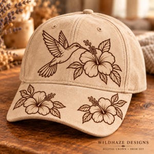 May include: A tan baseball cap featuring a dark brown line art illustration of a hummingbird hovering near a hibiscus flower on the crown, with two additional hibiscus flower illustrations on the brim. The text reads WILDHAZE DESIGNS.