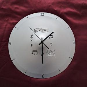 May include: A circular, silver clock featuring black hands and numerals. The clock face is decorated with musical notes and symbols, including a treble clef. The background is a deep burgundy colour.