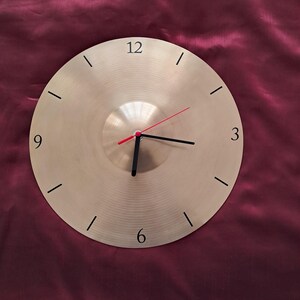 May include: A circular wall clock with a metallic gold finish. It features black hour markers and hands, with a red second hand. The clock face has a textured, concentric circle design. The numbers 12, 3, 6, and 9 are printed in black.