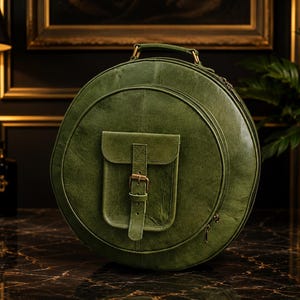 Könnte beinhalten: Eine runde olivgrüne Ledertasche mit einem Tragegriff und einer mittigen Außentasche, die mit einem vertikalen Riemen und einer Messingschnalle verschlossen wird. Die Tasche hat ein Reißverschluss-Hauptfach und eine glatte, leicht patinierte Oberfläche.
