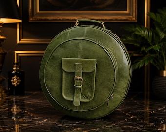 May include: A round olive green leather bag featuring a top handle and a central exterior pocket with a vertical strap and a brass buckle closure. The bag has a zippered main compartment and a smooth, slightly distressed finish on the leather surface.