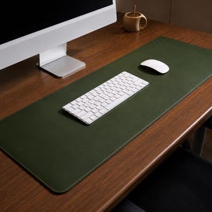 May include: A rectangular dark olive green desk mat with rounded corners and white perimeter stitching. The mat is positioned on a wooden surface, supporting a white wireless keyboard and a white wireless computer mouse for office use.