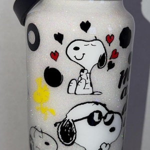 May include: A white glitter water bottle featuring Snoopy and Woodstock cartoon characters. The bottle has black accents, a black handle, and a black lid. The design includes black circles, red hearts, and the words "My Happy Place".