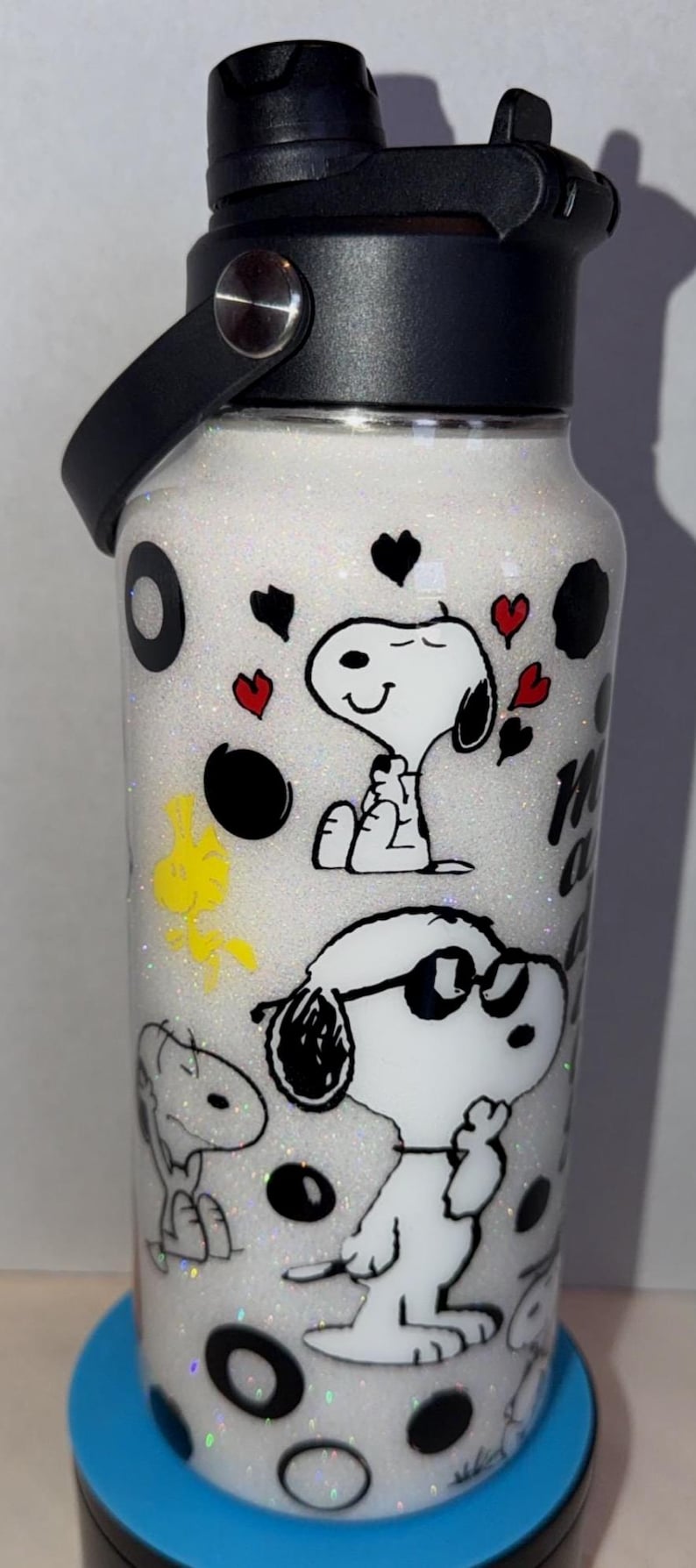 May include: A white glitter water bottle featuring Snoopy and Woodstock cartoon characters. The bottle has black accents, a black handle, and a black lid. The design includes black circles, red hearts, and the words "My Happy Place".