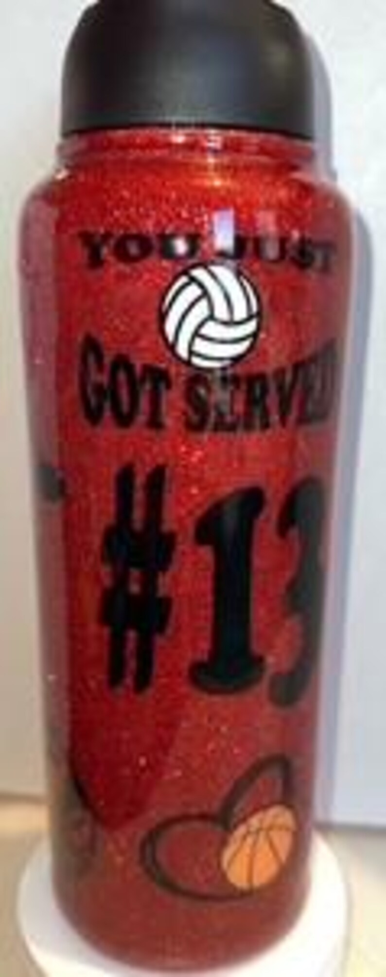 May include: Red glitter water bottle with a black lid. The bottle has the text "YOU JUST GOT SERVED" and the number "13" in black. A volleyball graphic and a basketball graphic are also featured.