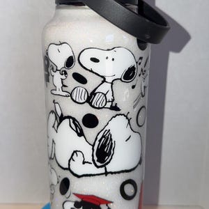 May include: White glitter water bottle featuring Snoopy and Woodstock cartoon characters in black and white. The bottle has a black lid and handle. The design includes Snoopy in various poses, with musical notes and black circles.