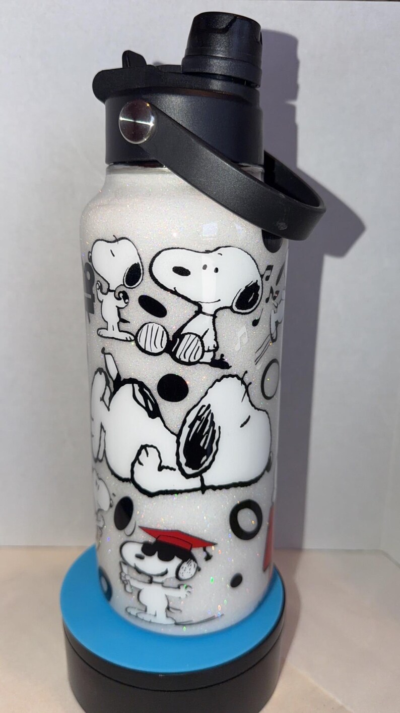 May include: White glitter water bottle featuring Snoopy and Woodstock cartoon characters in black and white. The bottle has a black lid and handle. The design includes Snoopy in various poses, with musical notes and black circles.