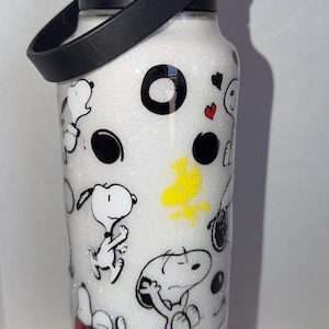May include: White water bottle with a black lid and handle, featuring a glittery finish and cartoon Snoopy and Woodstock designs. The bottle has a black handle and a red doghouse illustration.