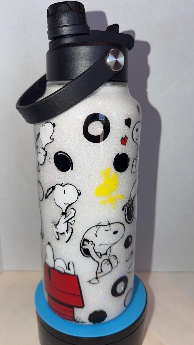 May include: White water bottle with a black lid and handle, featuring a glittery finish and cartoon Snoopy and Woodstock designs. The bottle has a black handle and a red doghouse illustration.