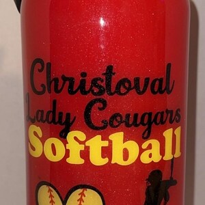 May include: Red glitter water bottle with black and yellow text that reads "Christoval Lady Cougars Softball." Includes a softball graphic and a silhouette of a softball player. The bottle has a black lid and handle.