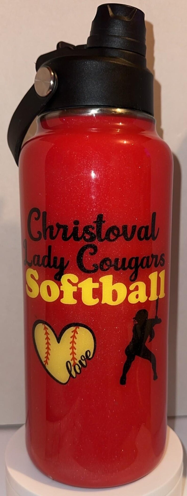 May include: Red glitter water bottle with black and yellow text that reads "Christoval Lady Cougars Softball." Includes a softball graphic and a silhouette of a softball player. The bottle has a black lid and handle.