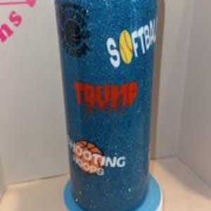 May include: A blue glitter water bottle with black accents. The bottle features the words "SOFTBALL", "TRUMP", and "SHOOTING HOOPS" in white and red lettering, along with a softball and basketball graphic. The bottle is sitting on a white and blue stand.