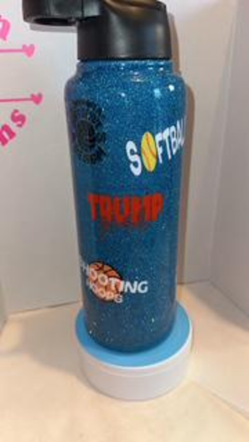 May include: A blue glitter water bottle with black accents. The bottle features the words "SOFTBALL", "TRUMP", and "SHOOTING HOOPS" in white and red lettering, along with a softball and basketball graphic. The bottle is sitting on a white and blue stand.