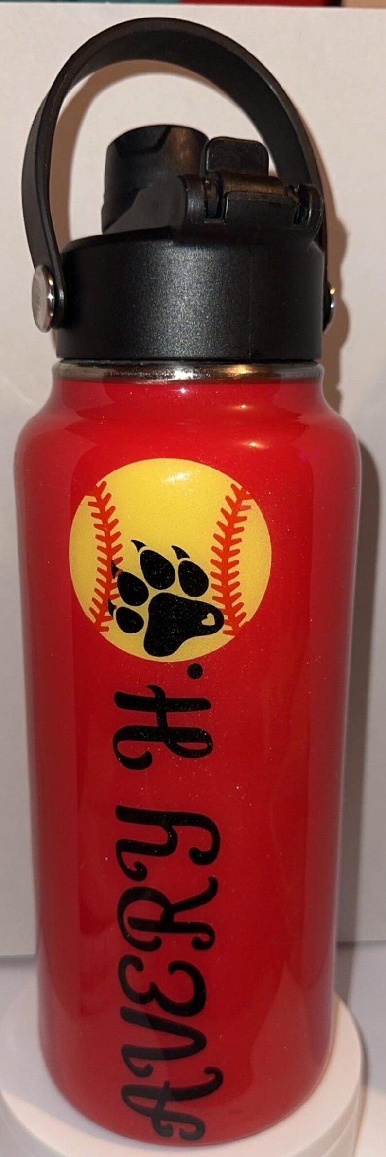 May include: Red water bottle with a black lid and handle. A yellow softball graphic with a black paw print is on the front. The name "Avery H." is written in black script.