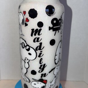 May include: A white glitter water bottle with a black lid and a black and white Snoopy design. The bottle has the name "Madie" in black script. The design includes Snoopy characters, hearts, and film reel icons.
