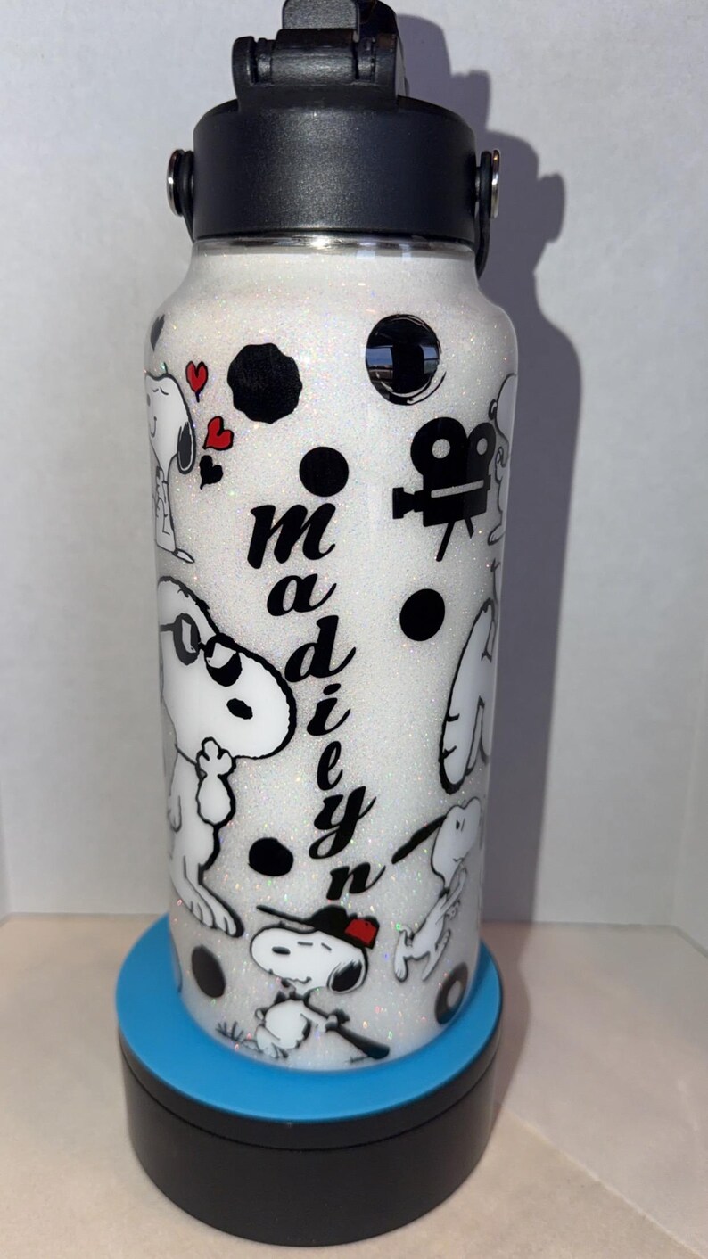 May include: A white glitter water bottle with a black lid and a black and white Snoopy design. The bottle has the name "Madie" in black script. The design includes Snoopy characters, hearts, and film reel icons.