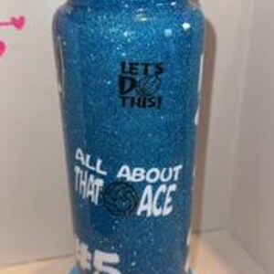 May include: A blue glitter water bottle with a black lid and a white base. The bottle has white text that reads "LET'S DO THIS!" and "ALL ABOUT THAT ACE #5". The bottle is approximately 9 inches tall.