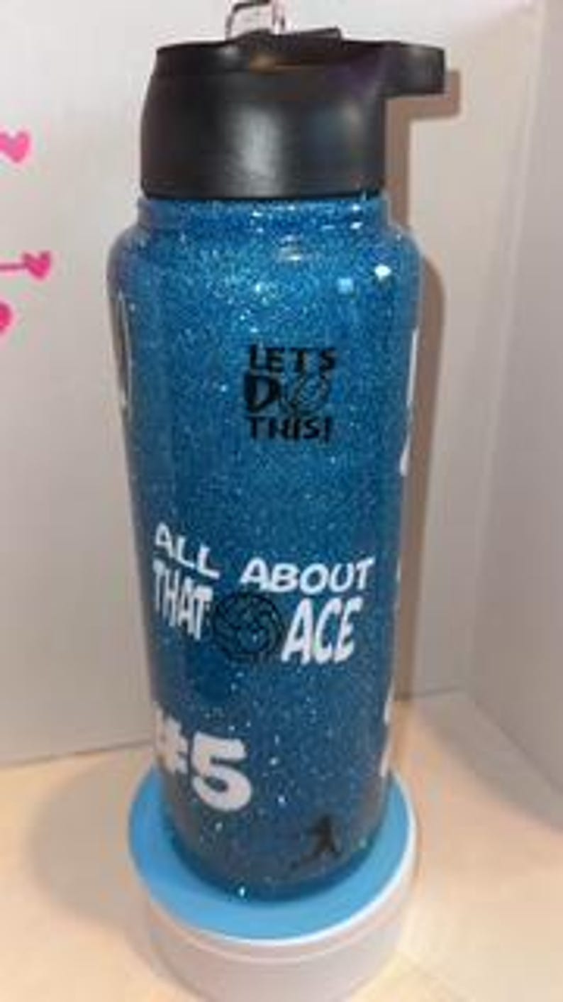 May include: A blue glitter water bottle with a black lid and a white base. The bottle has white text that reads "LET'S DO THIS!" and "ALL ABOUT THAT ACE #5". The bottle is approximately 9 inches tall.