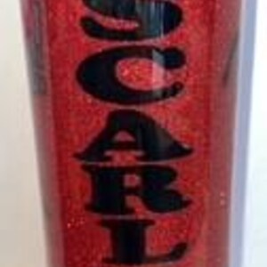 May include: A red glitter tumbler with the word "SCARLET" in black lettering. The tumbler has a black lid and a cylindrical shape. The glitter gives the tumbler a sparkling appearance.