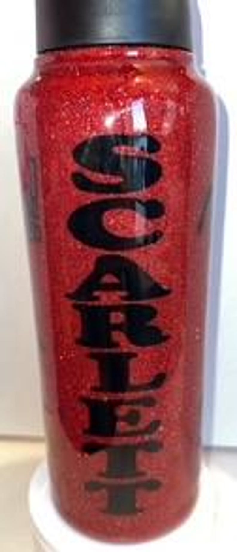 May include: A red glitter tumbler with the word "SCARLET" in black lettering. The tumbler has a black lid and a cylindrical shape. The glitter gives the tumbler a sparkling appearance.