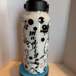 May include: White water bottle with a black lid and handle, decorated with cartoon characters and the name "Madiyn". The bottle features a black film camera, hearts, and polka dots. The bottle is sitting on a blue and black base.