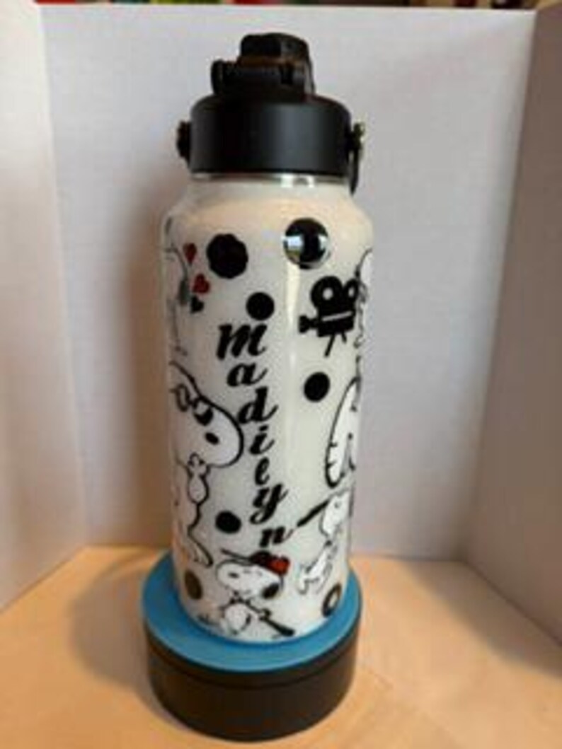 May include: White water bottle with a black lid and handle, decorated with cartoon characters and the name "Madiyn". The bottle features a black film camera, hearts, and polka dots. The bottle is sitting on a blue and black base.