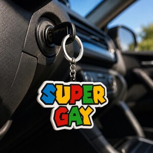 Può includere: Un portachiavi con un ciondolo multicolore dal bordo bianco che riporta la scritta SUPER GAY in un carattere a blocchi e pixelato. Le lettere sono colorate in blu, giallo, rosso e verde. Il ciondolo è fissato a un anello e catena in metallo.