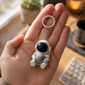 Może przedstawiać: Biały brelok w kształcie astronauty z błyszczącym czarnym wizjerem hełmu i małym, szarym, teksturowanym księżycem trzymanym w dłoniach figurki. Astronauta jest przymocowany do srebrnego metalowego łańcuszka i okrągłego kółka na klucze.