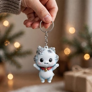 Può includere: Un portachiavi raffigurante una piccola creatura azzurra in stile cartone animato con grandi occhi neri, un collare rosso con un minuscolo campanellino dorato e accenti glitter argento sulla testa e sulle orecchie, fissato a un anello in metallo.