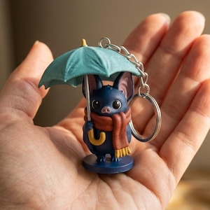 Op de afbeelding: Een kleine donkerblauwe vleermuis-sleutelhanger met een groenblauw parapluutje, een rode sjaal met gele franjes en een zilveren metalen ring. De vleermuis heeft grote donkere ogen, puntige oren en een kleine roze snuit op een ronde voet.