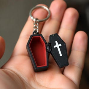 May include: A black coffin-shaped keychain with a hinged lid that opens to reveal a bright red interior. The exterior of the lid features a white cross emblem. The keychain is attached to a silver-toned metal split ring by a short chain.