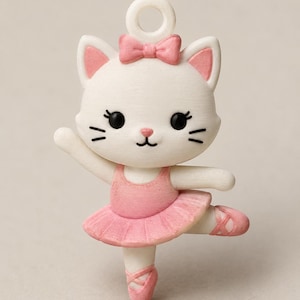May include: A white cat figurine in a ballet pose, wearing a pink tutu, leotard, and ballet slippers. The cat has a pink bow on its head and black eyes. The figurine is designed with a loop for hanging.