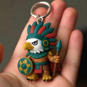 May include: A keychain featuring a stylized eagle warrior figure wearing a teal and red feathered headdress. The figure has a yellow beak, holds a teal and yellow circular shield in one hand, and a brown spear in the other, with a metal ring attached.