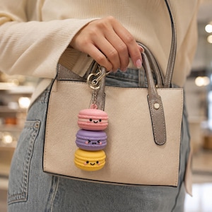 May include: A keyring featuring three stacked macaron charms in pink, purple, and yellow, each with a unique printed facial expression. The pink macaron is on top, the purple one is in the middle, and the yellow one is at the bottom.