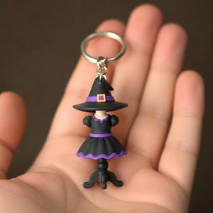 Puede incluir: Un llavero que presenta un vestido negro en miniatura sobre un soporte, rematado con un sombrero de bruja negro. El vestido tiene una cintura morada y un dobladillo con borde morado. El sombrero incluye una banda morada y una hebilla cuadrada amarilla.