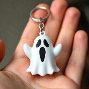 Może przedstawiać: Brelok w kształcie białego ducha ze srebrnym kółkiem. Duch ma czarne oczy i usta. Ten brelok to zabawny dodatek, idealny na Halloween lub do codziennego użytku.