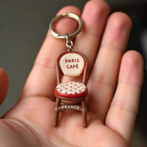 May include: A miniature brown chair keyring featuring a cream-coloured backrest with the red text PARIS CAFÉ, a cream seat with red polka dots, and the word FRANCE printed in white on the front horizontal support bar of the chair frame.