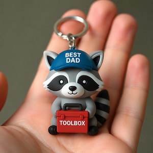 Puede incluir: Un llavero con un personaje de mapache gris y blanco que lleva una gorra de béisbol azul con el texto blanco BEST DAD impreso. El mapache sostiene una pequeña caja de herramientas rectangular roja con el texto TOOLBOX en blanco al frente.