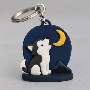 May include: A circular keychain featuring a dark blue background with a yellow crescent moon and black mountain silhouettes. A white and black husky puppy is depicted in the center, sitting and looking upward with closed eyes.