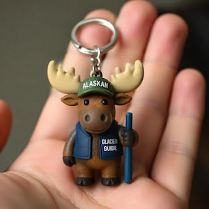 May include: A keychain featuring a brown moose figurine wearing a green cap with the text ALASKAN and a blue vest with the text GLACIER GUIDE. The moose holds a blue staff in one hand and is attached to a silver metal split ring by a chain.