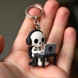Può includere: Un portachiavi con una piccola figura di scheletro bianco che indossa cuffie nere, seduta su una sedia da ufficio nera a una scrivania nera mentre usa un portatile grigio con una piccola icona di teschio nero sul coperchio, fissato a un anello.