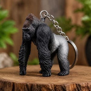 Puede incluir: Un llavero con una figura tridimensional detallada de un gorila de espalda plateada. El gorila se muestra en una postura cuadrúpeda, con pelaje negro texturizado en la cabeza y los brazos, que se vuelve de un tono gris claro en la espalda.