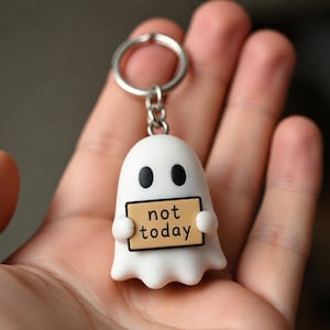 May include: A white ghost keychain featuring two black oval eyes and holding a small tan rectangular sign with the black text "not today" printed on it. The ghost is attached to a silver metal split ring by a short silver metal chain link.