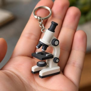Może przedstawiać: Brelok w kształcie miniaturowego mikroskopu z białą ramą i czarnymi akcentami, w tym okularem, obiektywami, stolikiem przedmiotowym i pokrętłami regulacyjnymi. Urządzenie jest przymocowane do srebrnego metalowego kółka za pomocą krótkiego łańcuszka.