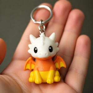 May include: A keychain featuring a small, stylized dragon figurine with a white head, orange torso, and yellow lower body and feet. The dragon has small orange wings with yellow accents and large black eyes. It is attached to a silver metal ring.